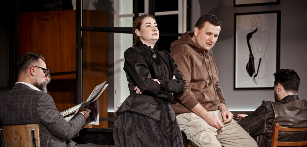 Scene from Jenůfa at Janáček Brno Festival. A man reads a newspaper at a table, two people lean against the table, and another man sits at the other end of the table.