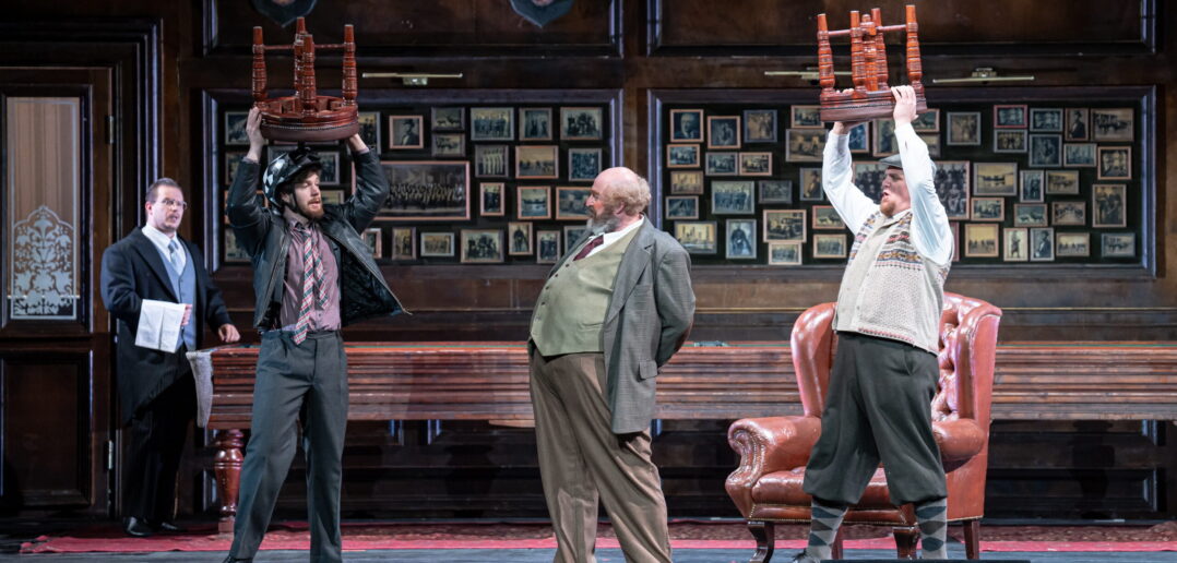 Four men on stage: two hold stools over their heads while looking at the protagonist, Falstaff, who stands in between them.