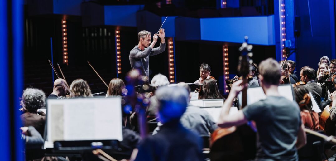 Alexander Shelley and Canada's NAC Orchestra in the middle of a performance of Strauss against a blue background.