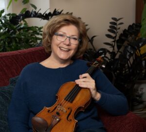 Gwen Hoebig smiling while holding a violin on a couch.